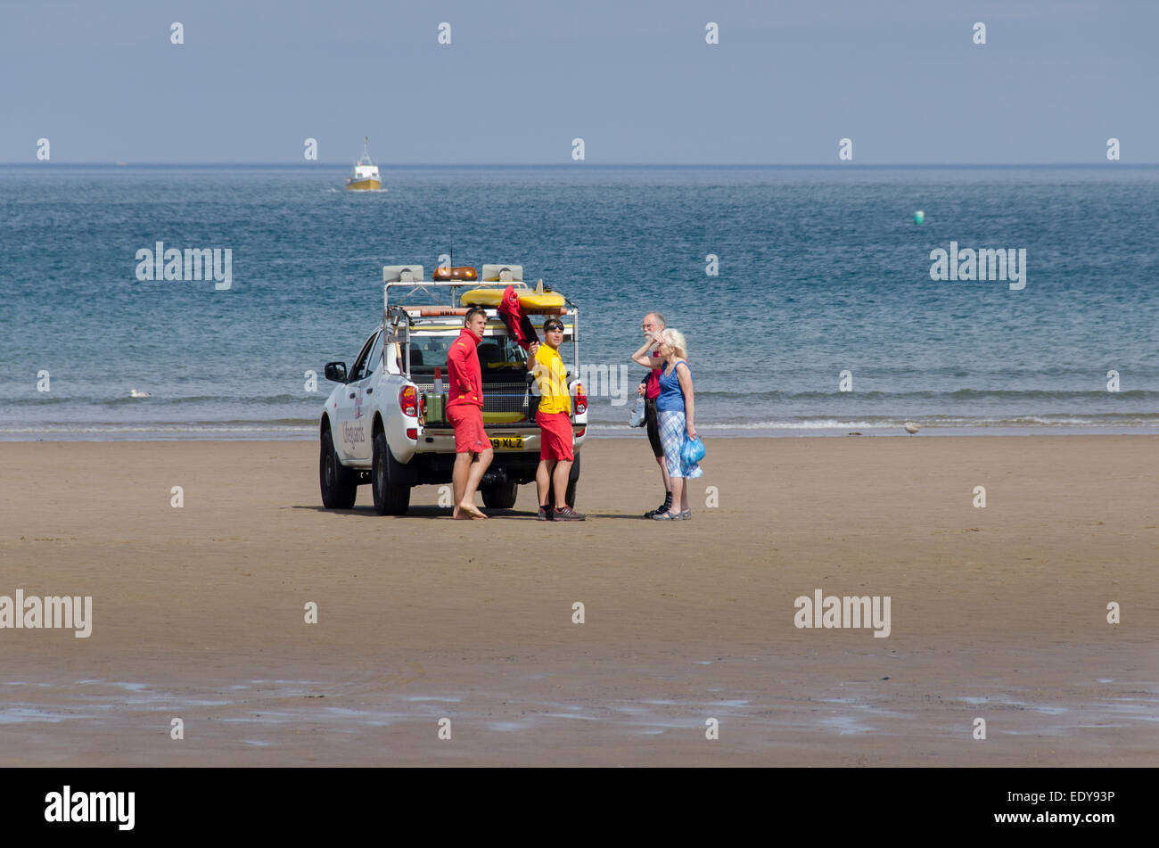2 male RNLI lifeguards standing by 4x4 patrol vehicle talking & giving advice to a couple of holidaymakers - Whitby beach, North Yorkshire, England. Stock Photo