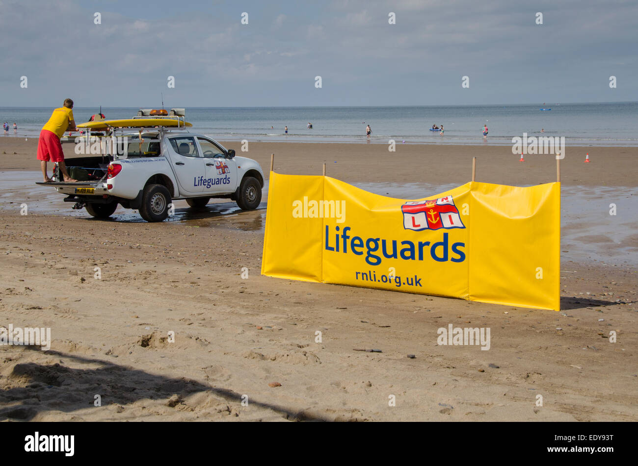 Young man on duty as RNLI lifeguard is watching people in sea & standing on back of parked pick-up truck - Whitby beach, North Yorkshire, England, UK. Stock Photo