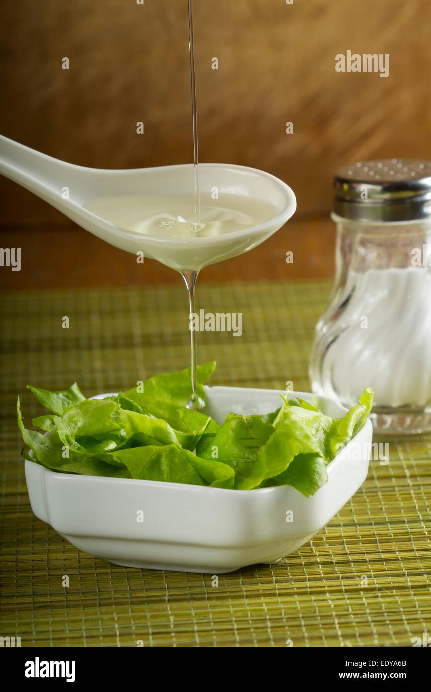Pouring oil over fresh salad in white bowl over green bamboo rug Stock Photo