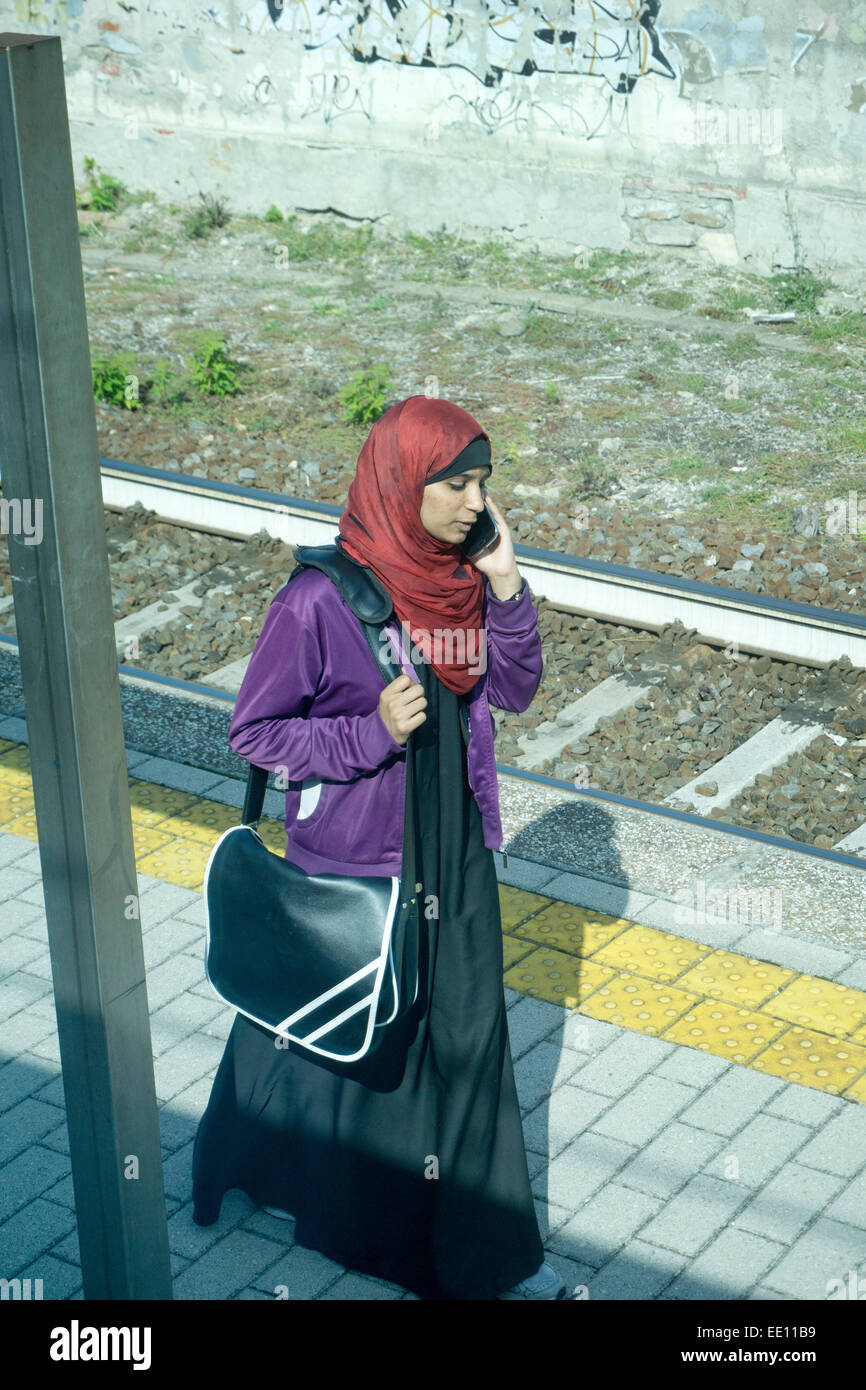 young Muslim woman with cellphone wears richly colored head scarf & jacket with large plastic satchel on suburban train platform Stock Photo