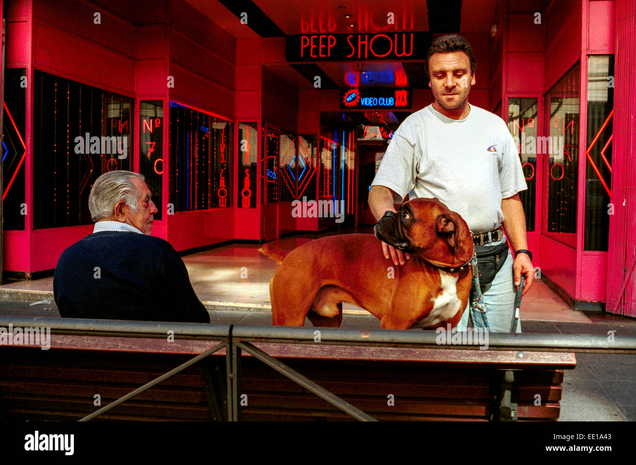 Man with a muzzled Boxer dog in front of a Peep Show in Madrid, Spain, next to a senior man on a bench Stock Photo