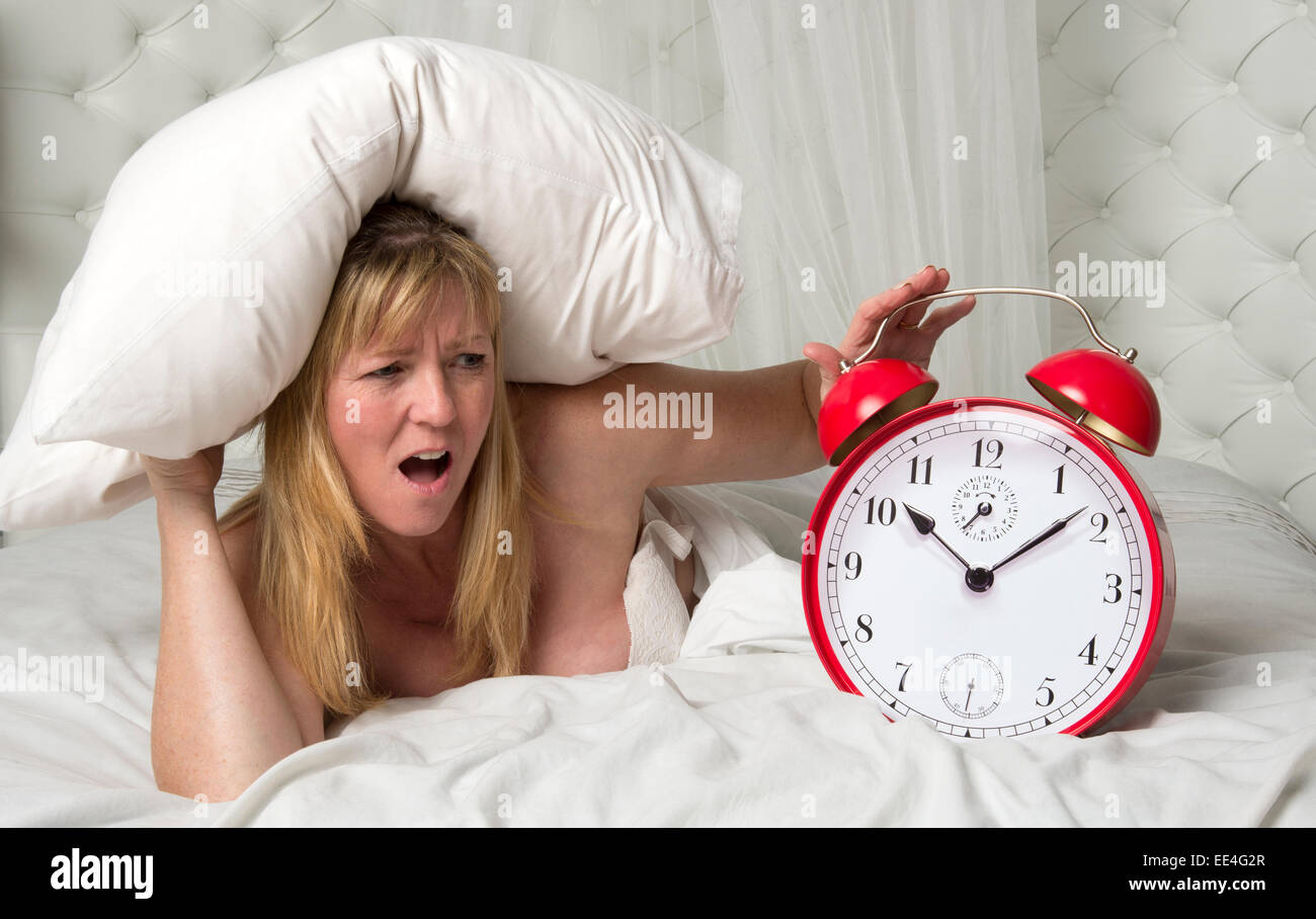 Woman in bed and getting angry with ringing alarm clock holds a pillow over her ears to deaden the noise Stock Photo