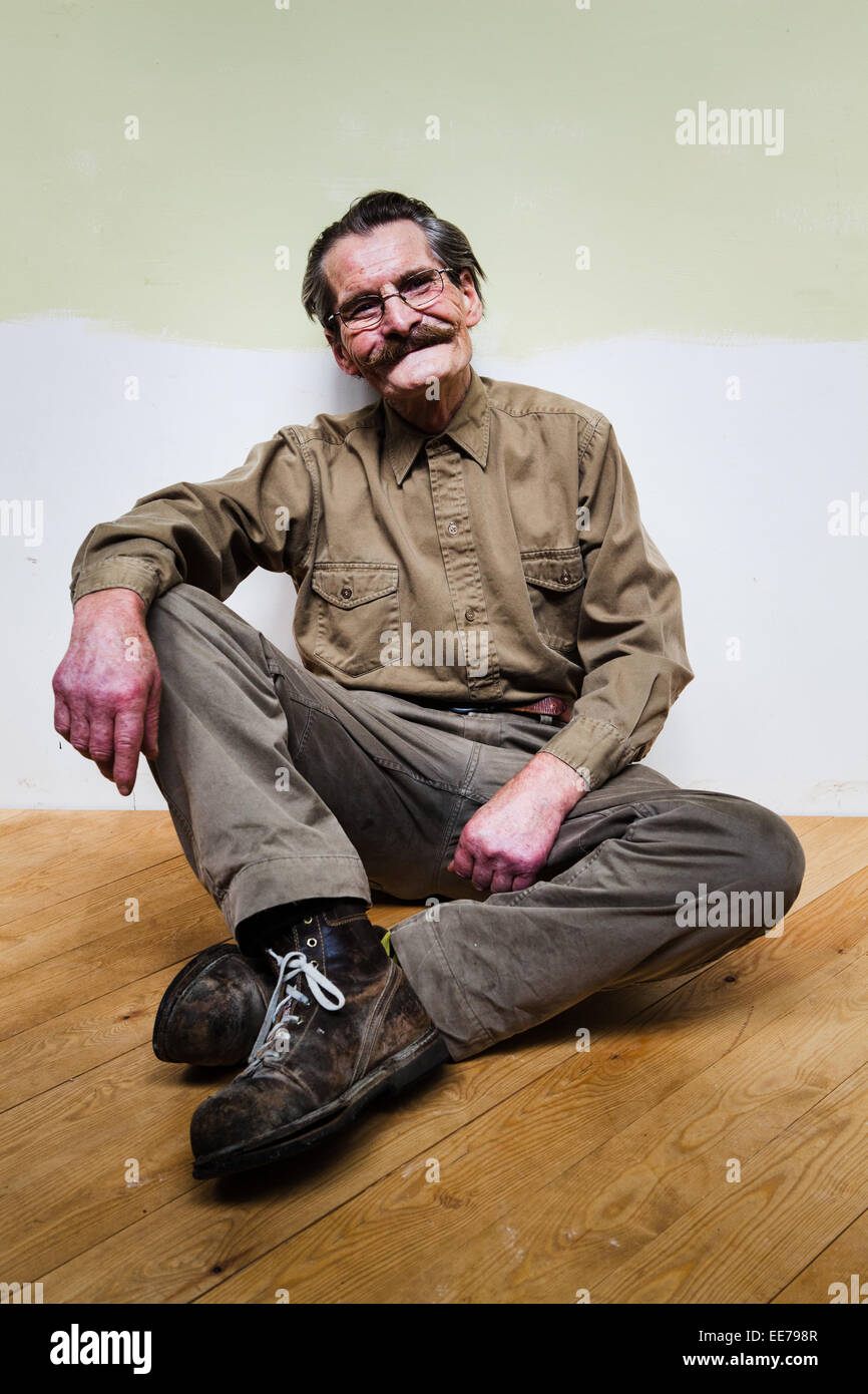 Man in his 60s with moustache wearing khaki shirt and trousers sitting on the floor. Stock Photo