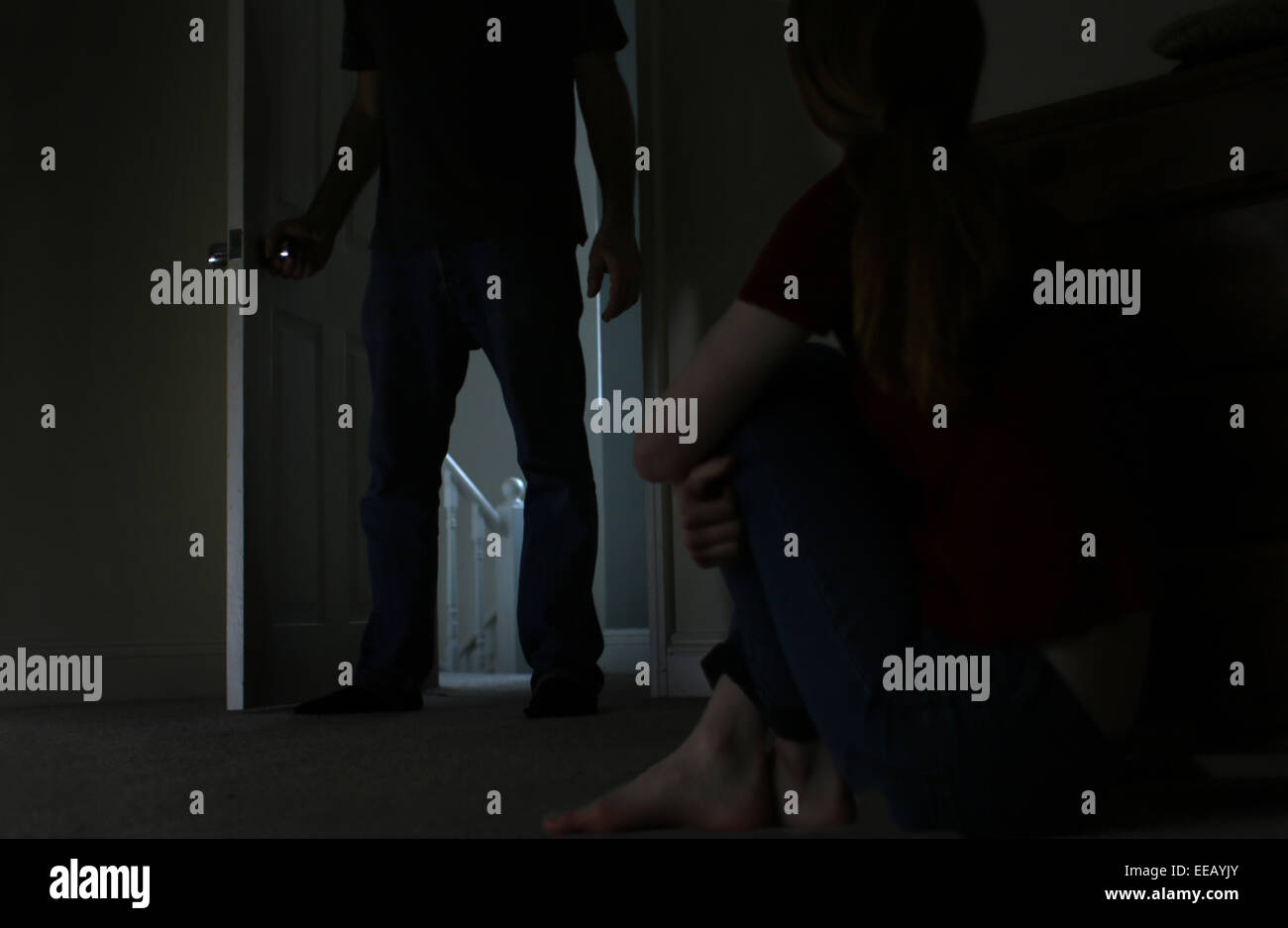Silhouette of a man, lower body shot, entering a dark room, silhouette of a teenage girl sitting cross legged on the floor. Stock Photo
