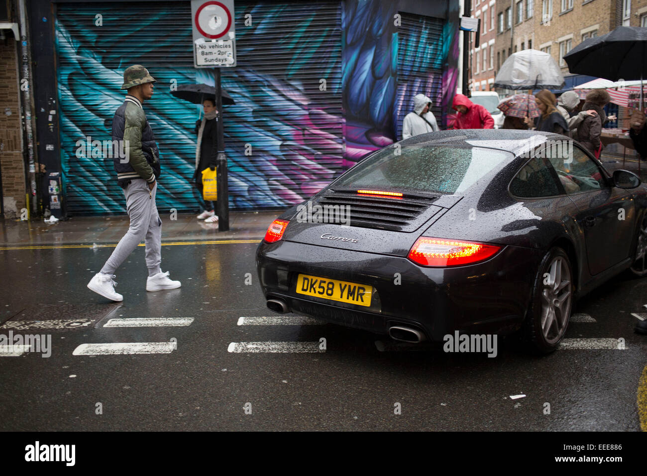 Wet day on Brick Lane in the East End of London, UK. Porche 911 drives through the market as people go about their day. It is an illustration of the haves and have nots as a sports car worth tens of thousands of pounds drives past stalls selling super cheap goods. Stock Photo