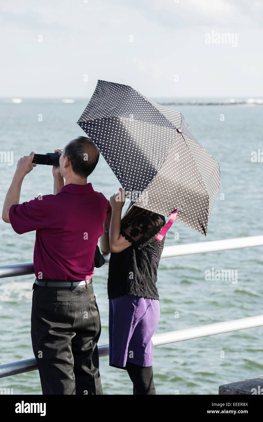 Back view of older Asian man with camcorder and woman hidden under a polk-a-dot umbrella standing at a railing looking at the Atlantic ocean. Stock Photo