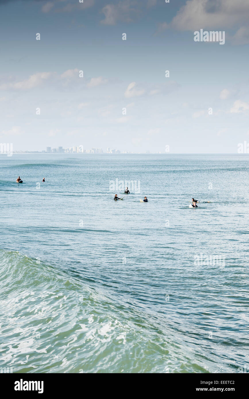 Seven surfers line up on their surfboards on a sunny day waiting to catch a wave as a swell rolls by with the Daytona Beach, FL skyline in background. Stock Photo