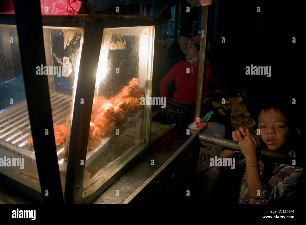 A young boy is leaning on a food cart containing fried chicken on a city street in Kampong Cham, Cambodia. Stock Photo