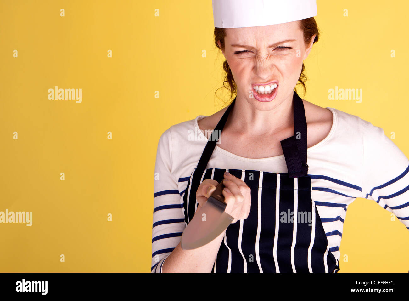 A young and angry female chef pointing a chefs knife on a yellow