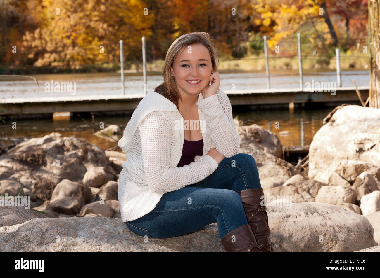 Young adult female smiling for portrait and sitting on boulder by lake Stock Photo