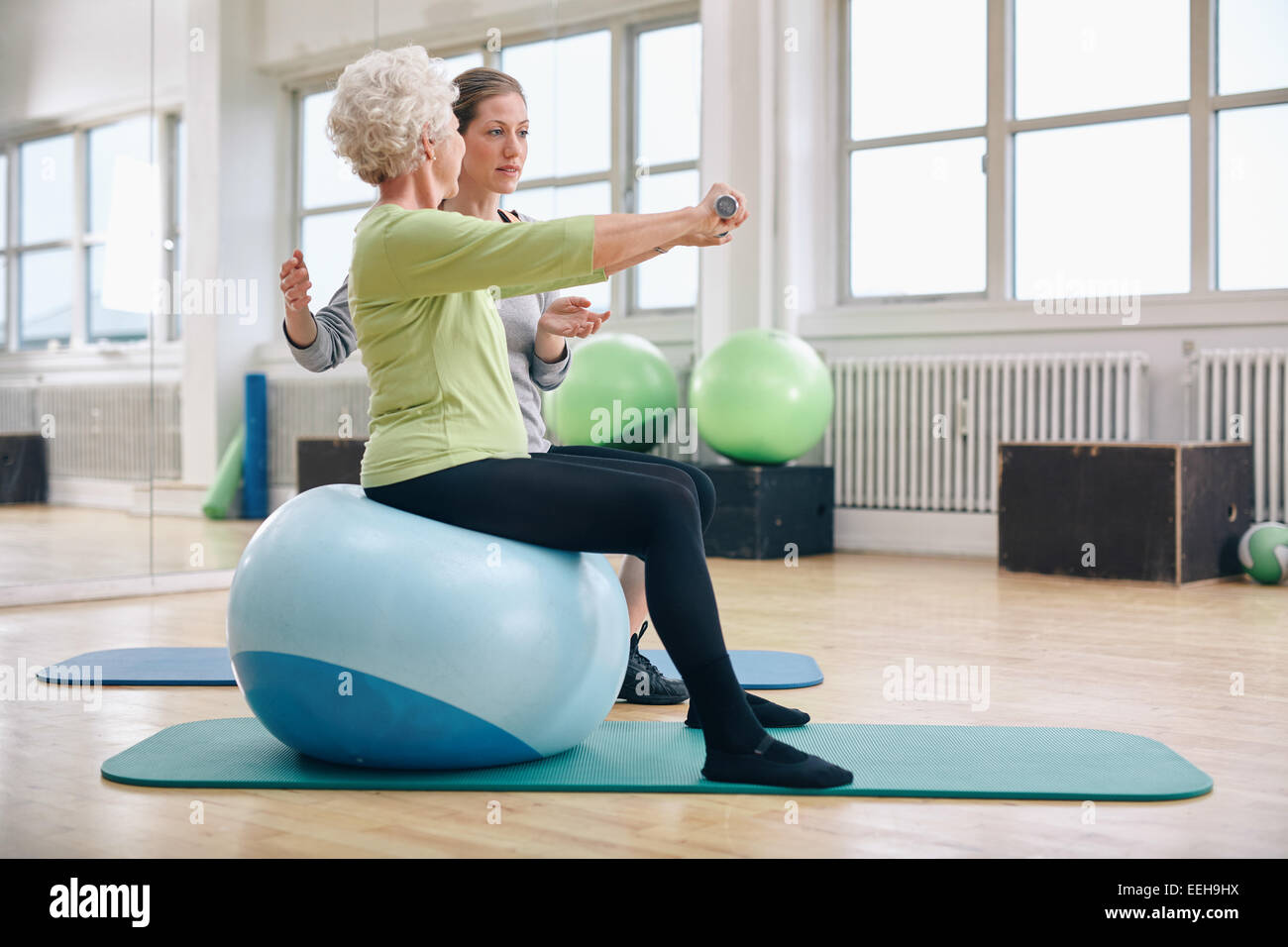 Female trainer assisting senior woman lifting weights in gym. Senior woman sitting on pilates ball doing weight exercise. Stock Photo