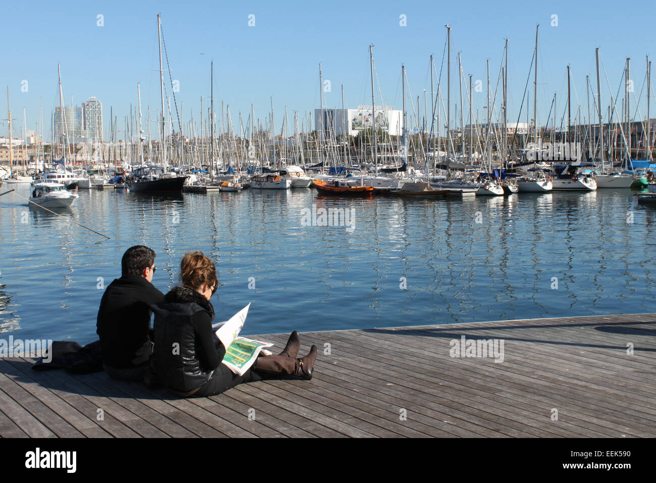 Couple sitting outside in the sunshine, on walkway of marina, Maremagnum, Barcelona on a cold winter's day Stock Photo