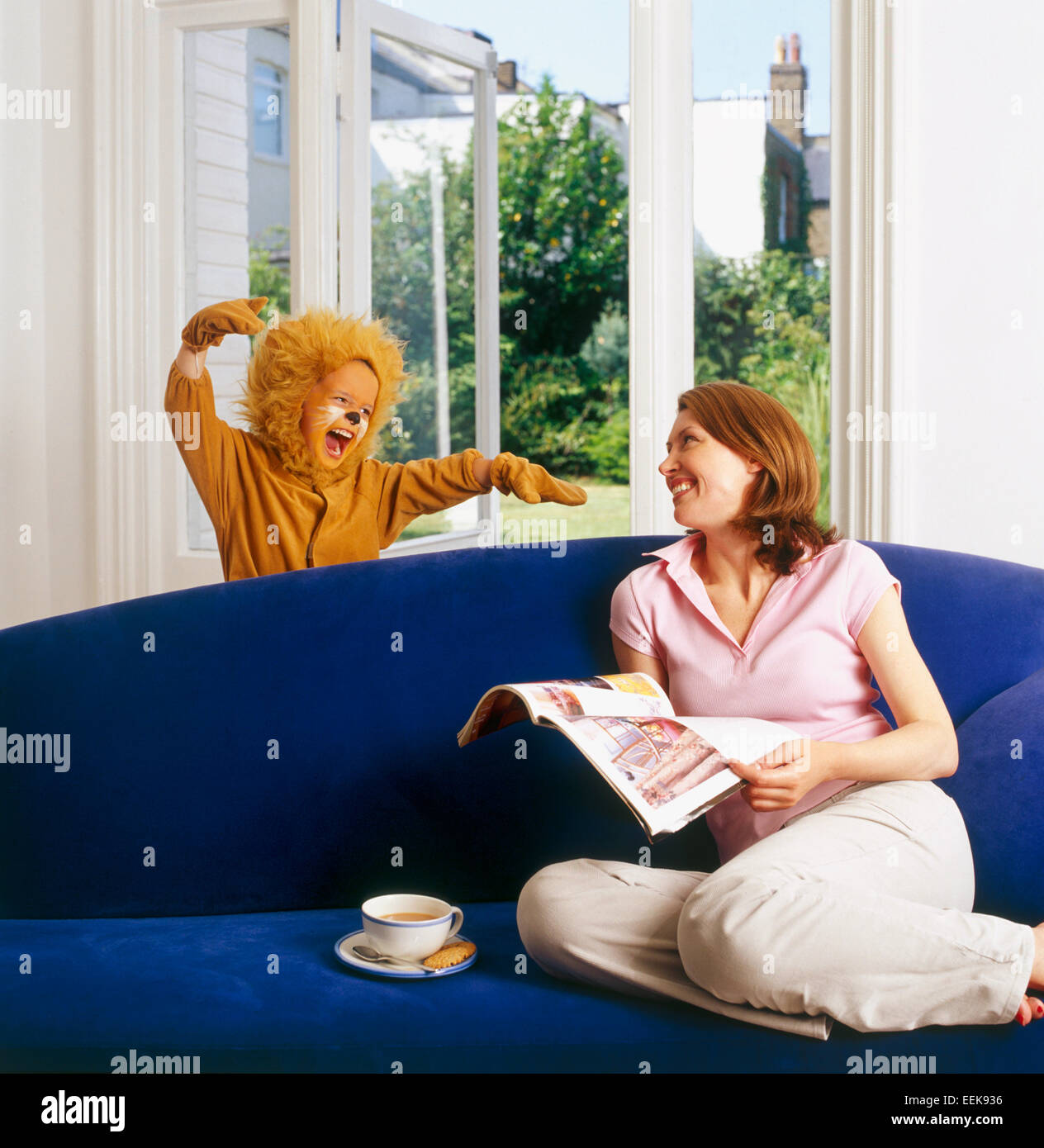 Mum at home sitting relaxing, reading a newspaper, her child dressed as lion surprises her from behind the sofa, roaring scarily Stock Photo