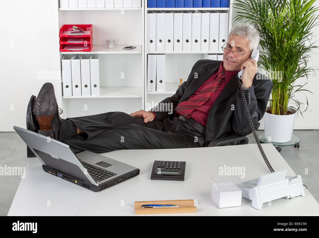 Mann mittleren Alters sitzt im Büro hat lässig die Beine auf dem Schreibtisch und telefoniert, Middle-aged man sits in an office Stock Photo