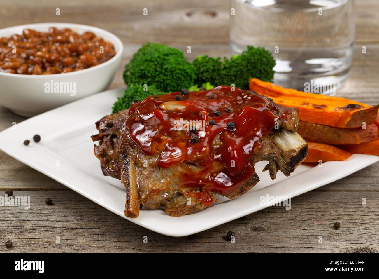 Close up of barbecued spare ribs with yam fries, broccoli, baked beans and glass of water on rustic wooden table. Stock Photo