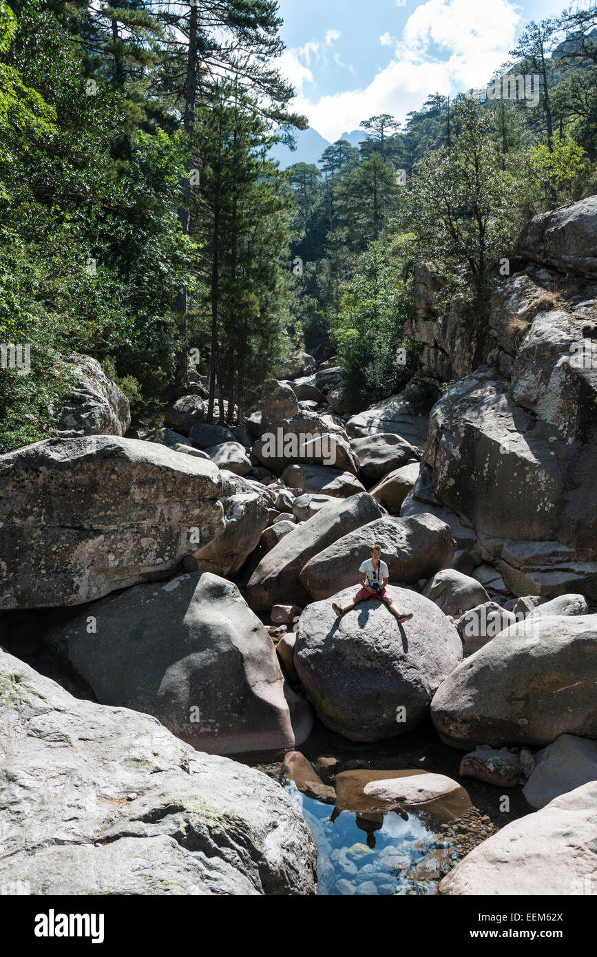 Young man sitting on rocks at the Calenzana river, mountains, hiking trail to the Refuge de Carrozzu Stock Photo