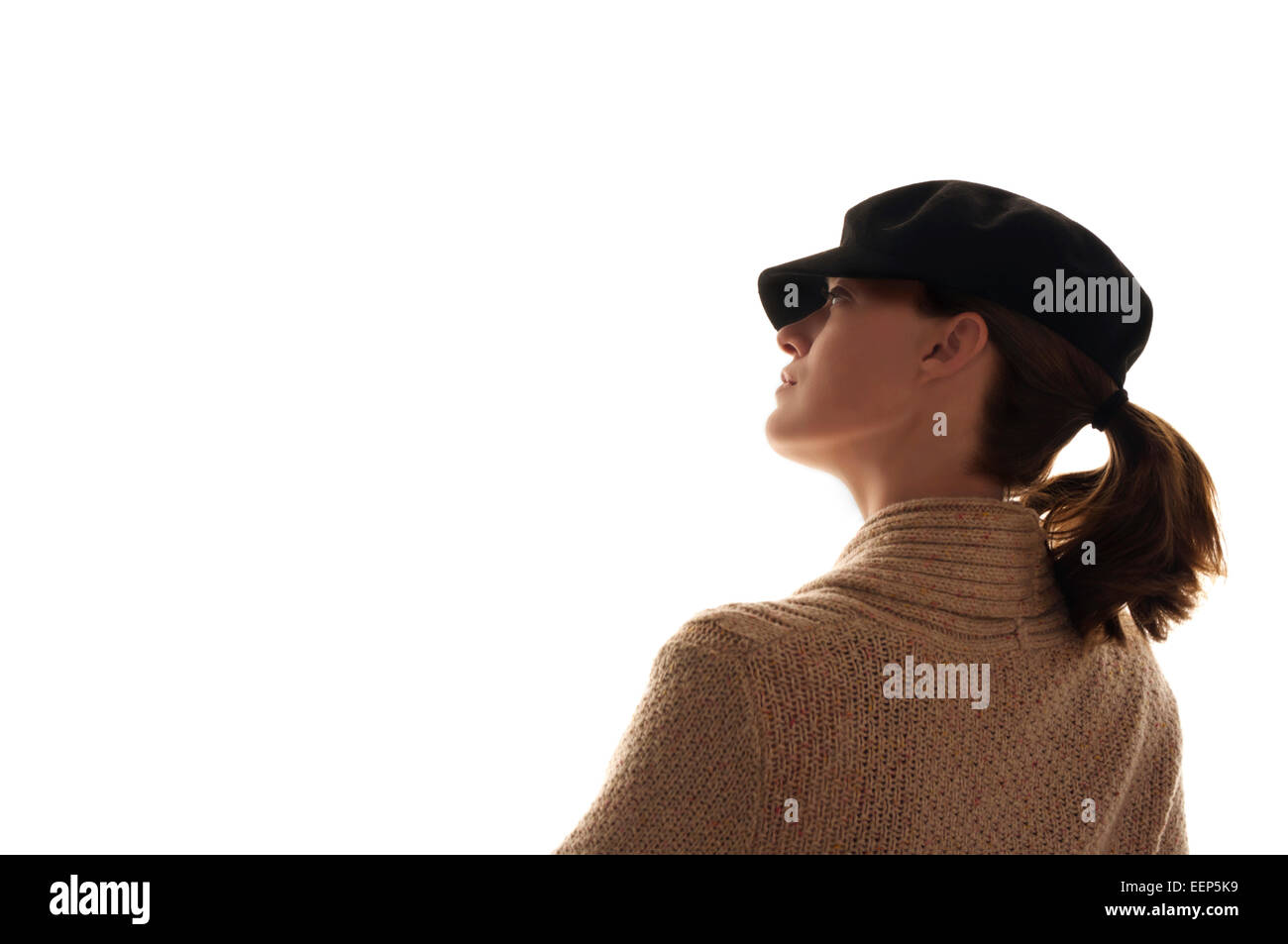 Side profile of a womans face looking upwards off in the distance she wears a black hat, hair in ponytail and a tan sweater. Stock Photo