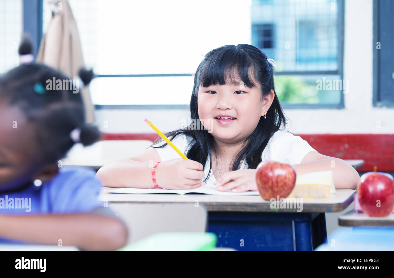 Asian girl smiling at classroom desk drawing on her book during a school lesson. Stock Photo
