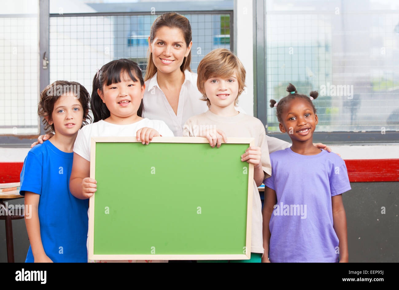 Happy multi-ethnic elementary classroom holding empty green chalkboard. Stock Photo