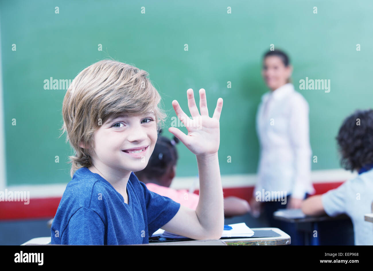 Happy little schoolboy raising hand while sitting at desk with ...