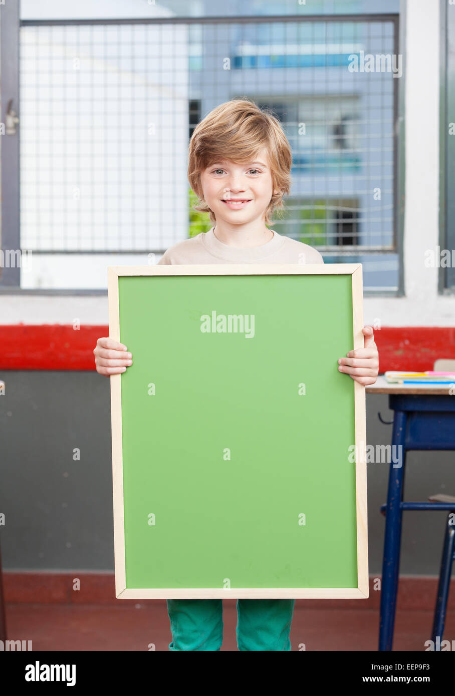 Smiling kid at primary school holding empty green chalkboard. Stock Photo