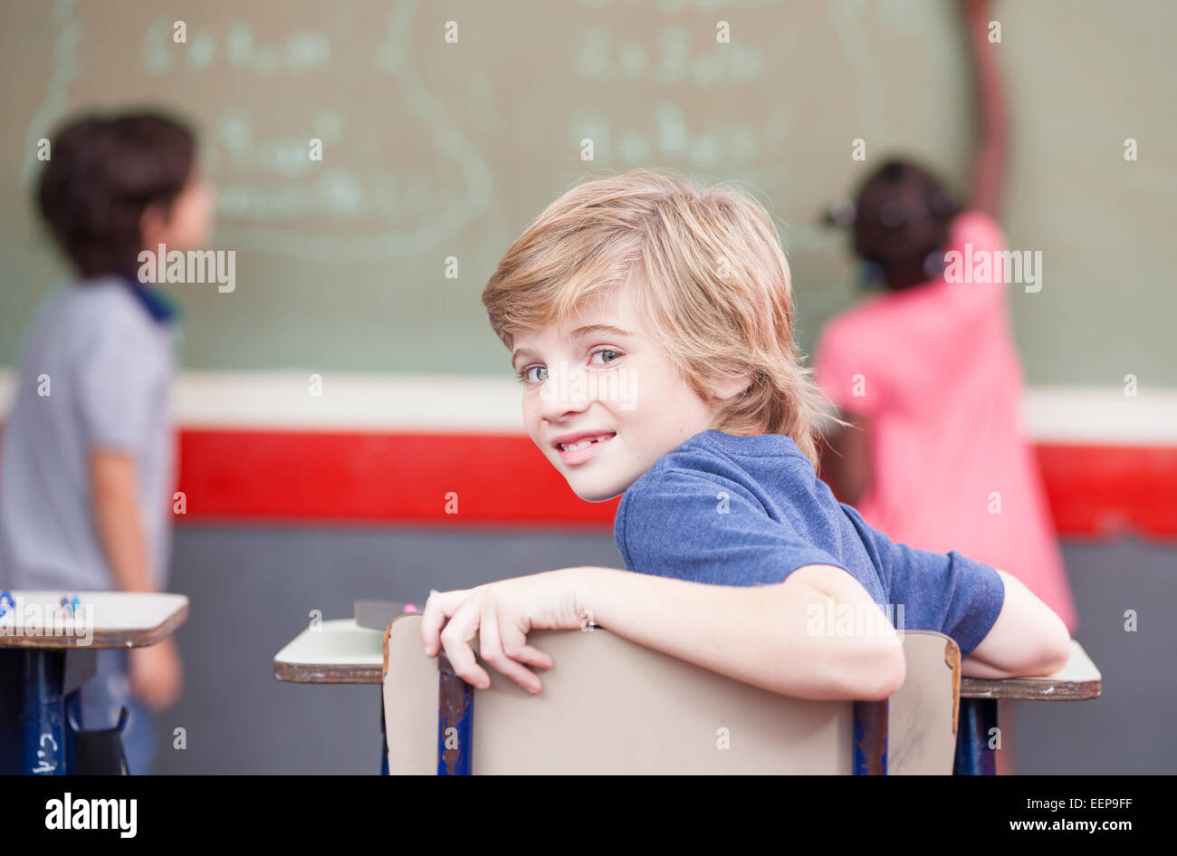 Multi ethnic elementary classroom. Kid looking at camera while classmates at chalkboard. Stock Photo