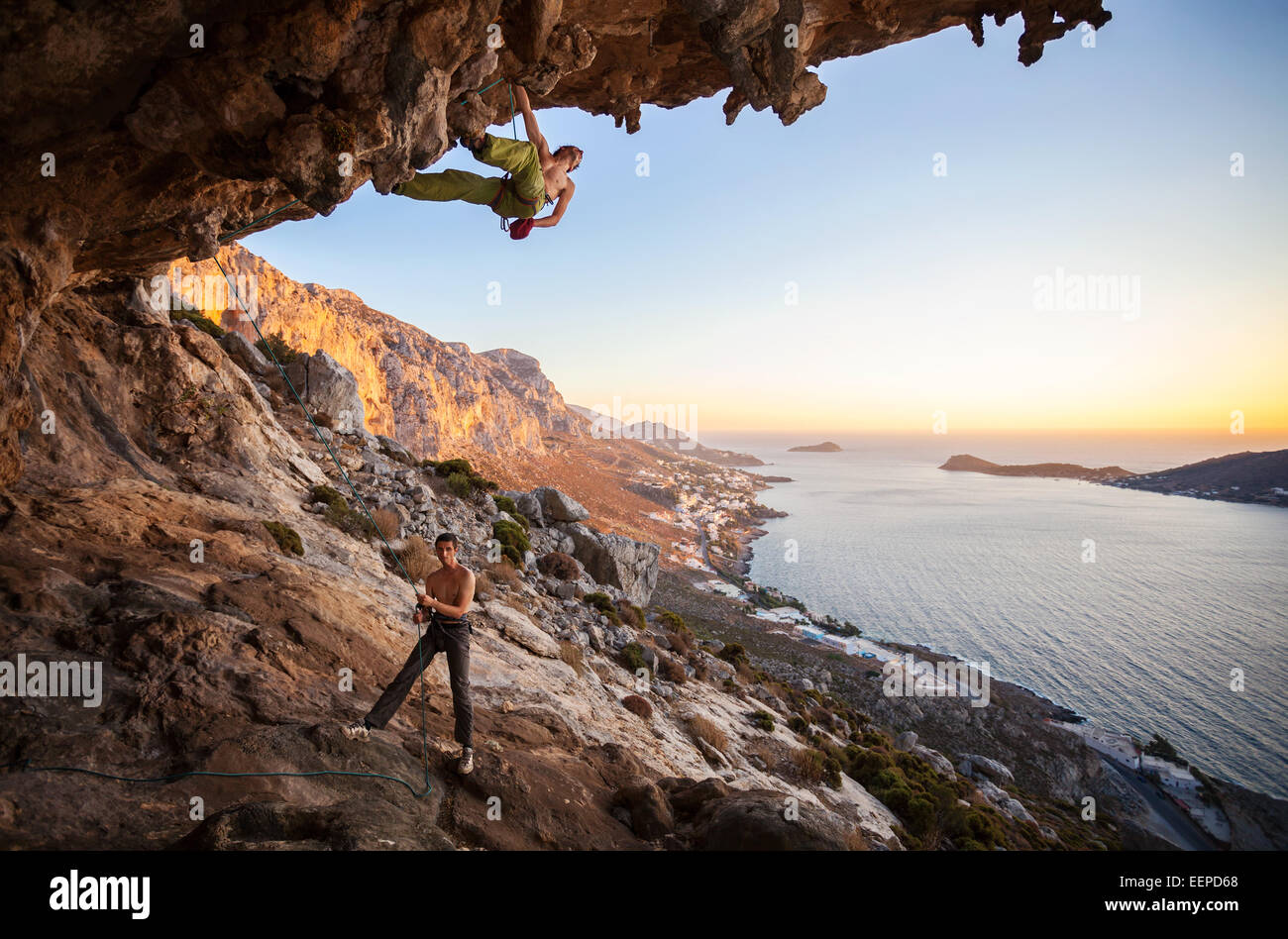 Male rock climber climbing on a roof in a cave, his partner belaying