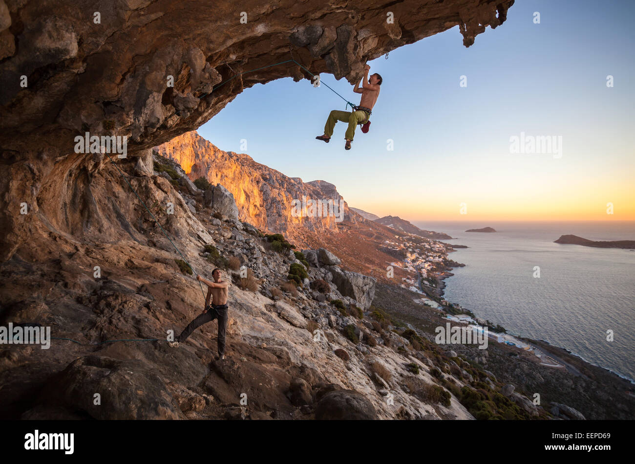 Male rock climber climbing on a roof in a cave, his partner belaying