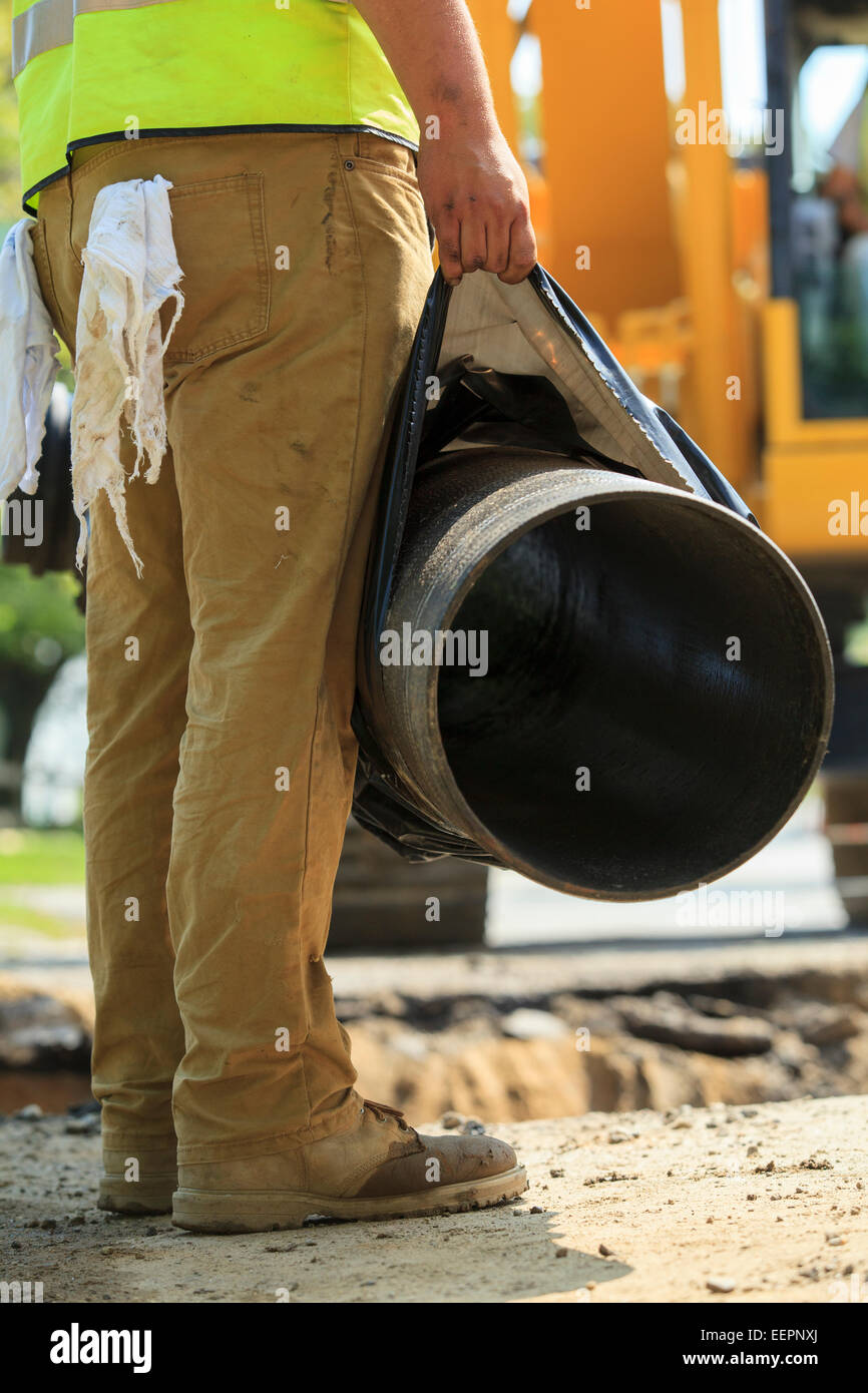 Construction worker holding water main pipe suspended by excavator Stock Photo