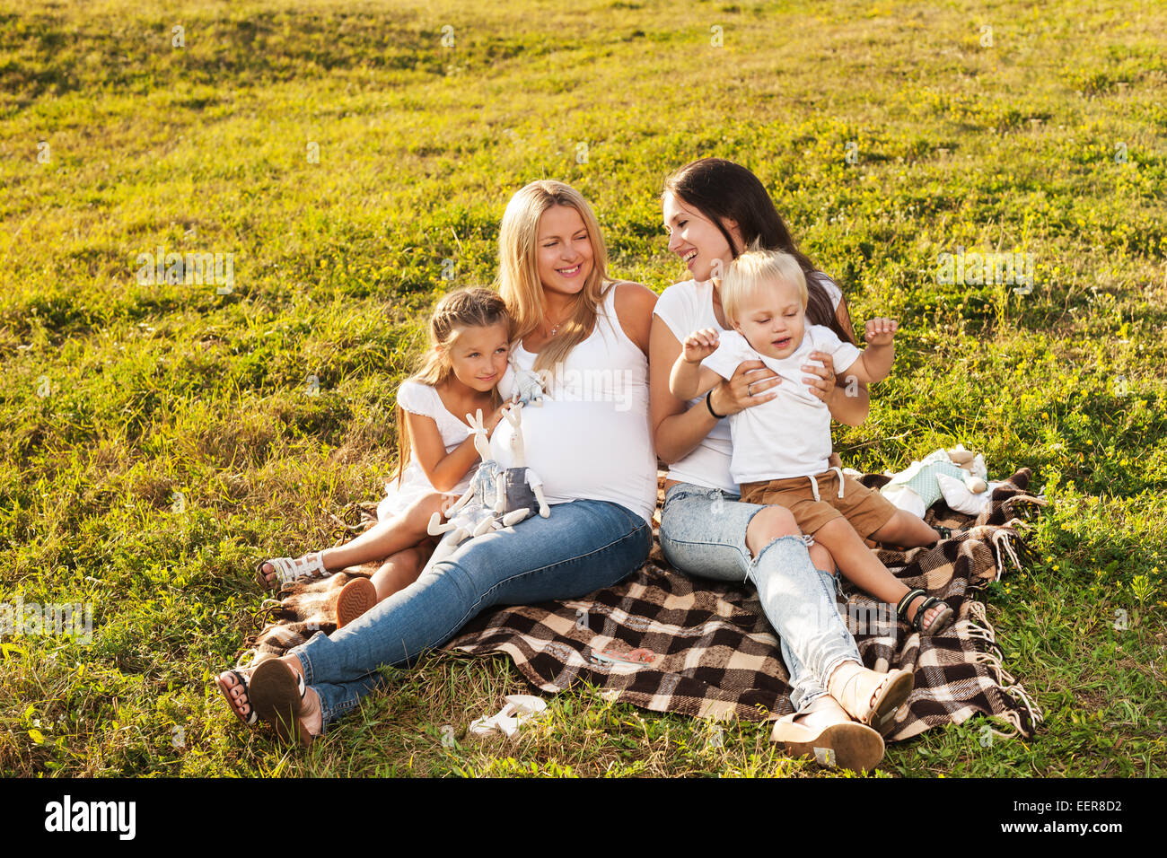 Two young women with kids enjoying sunshine in park. Beautiful pregnant woman with her friend and their kids. Mother's day conce Stock Photo