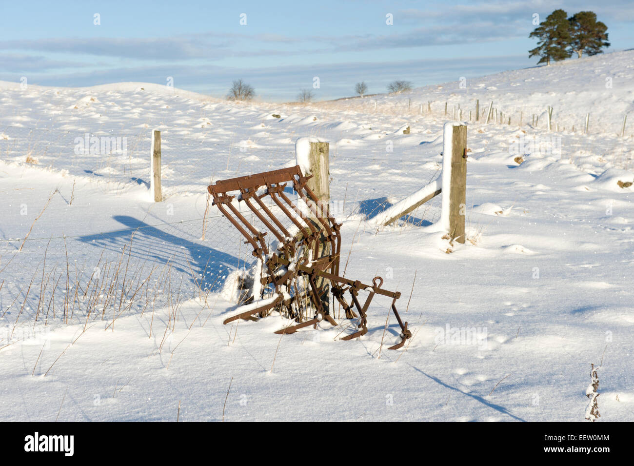 A rusty and abandoned chain harrow against a fence at the edge of a winter field covered in snow Stock Photo