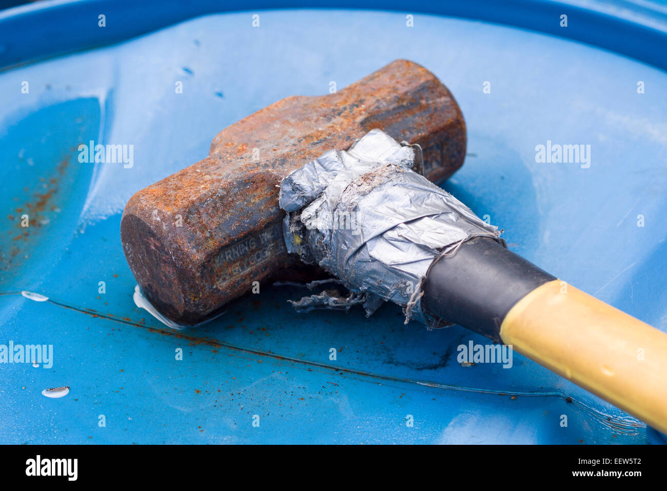 Rusty Sledge Hammer fixed with duct tape. A rusted sledge rests on a blue plastic barrel. Its handle repaired w duct tape Stock Photo