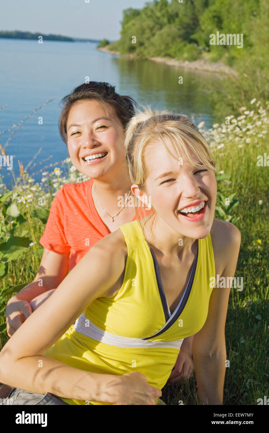 Two girl friends sitting and laughing in front of grass and water Stock Photo