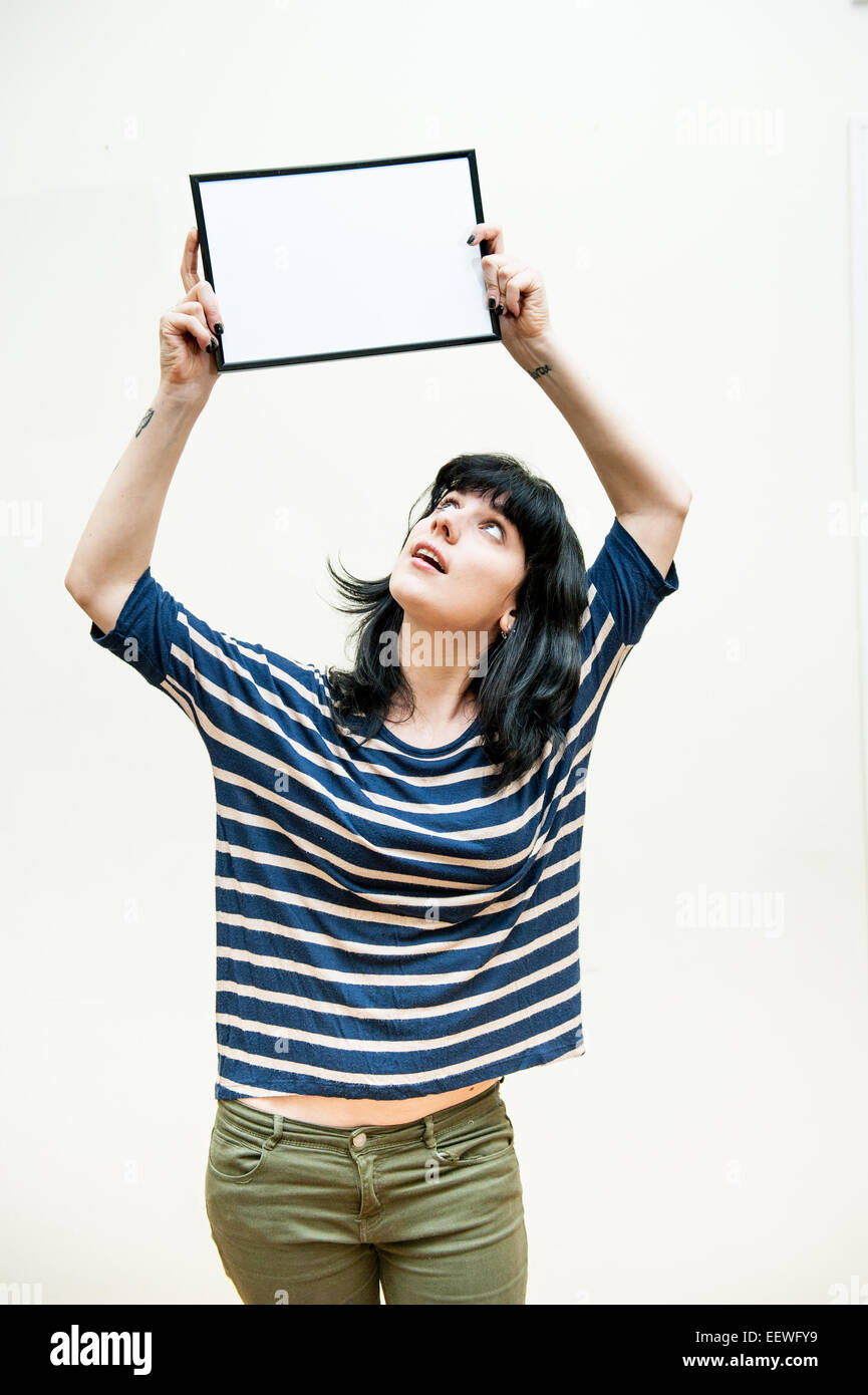 Pretty brunette girl looking and showing blank picture frame up on her head on white background Stock Photo