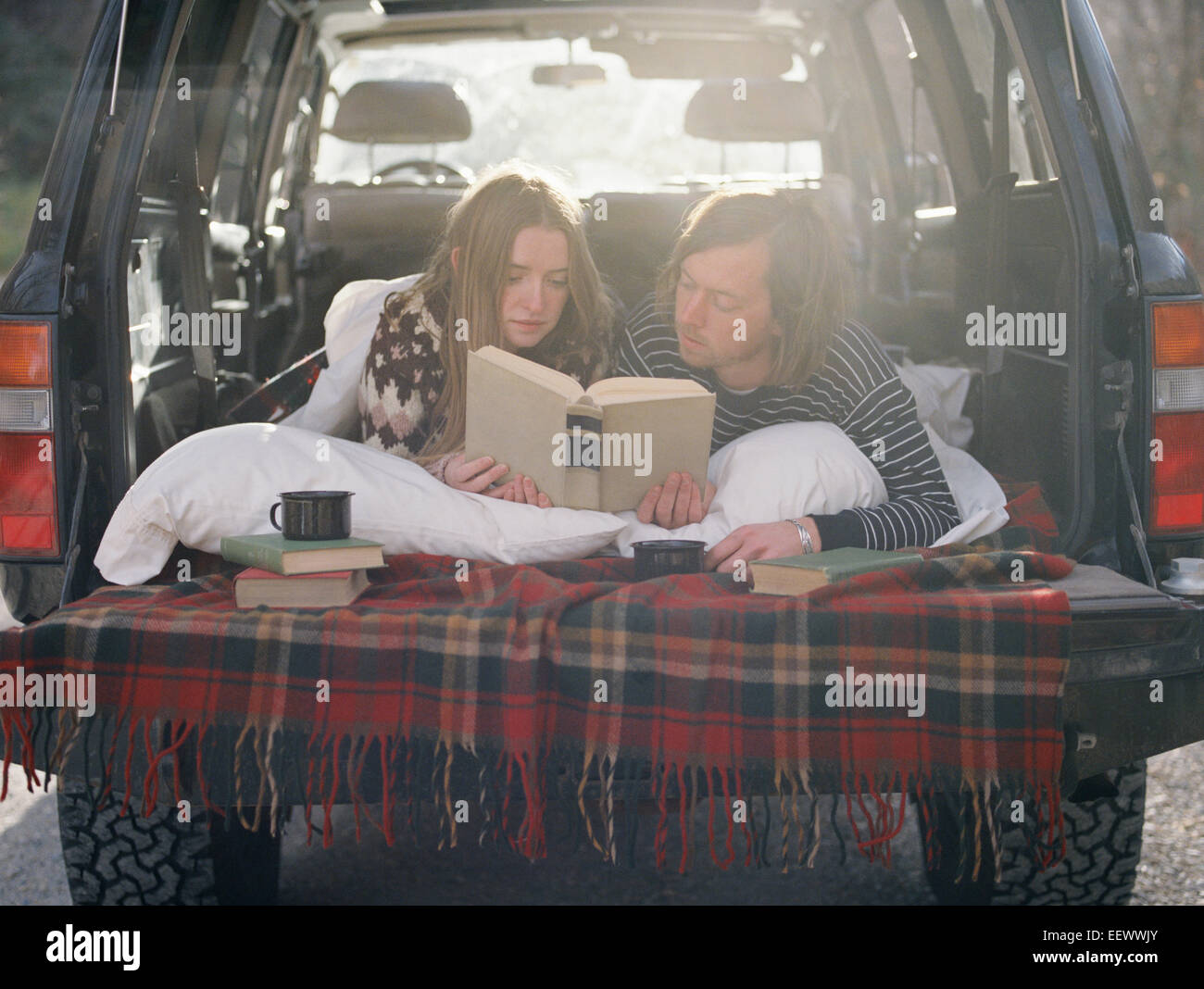 Young couple lying in the back of their car, reading a book. Stock Photo