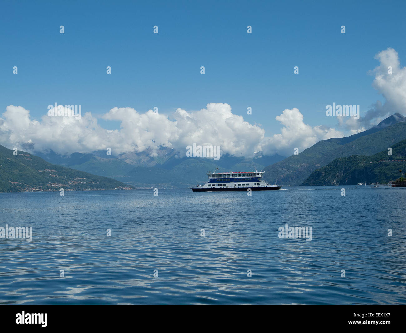 A ferry crosses Lake Como in Italy with a mountain backdrop tipped by clouds and a blue sky Stock Photo