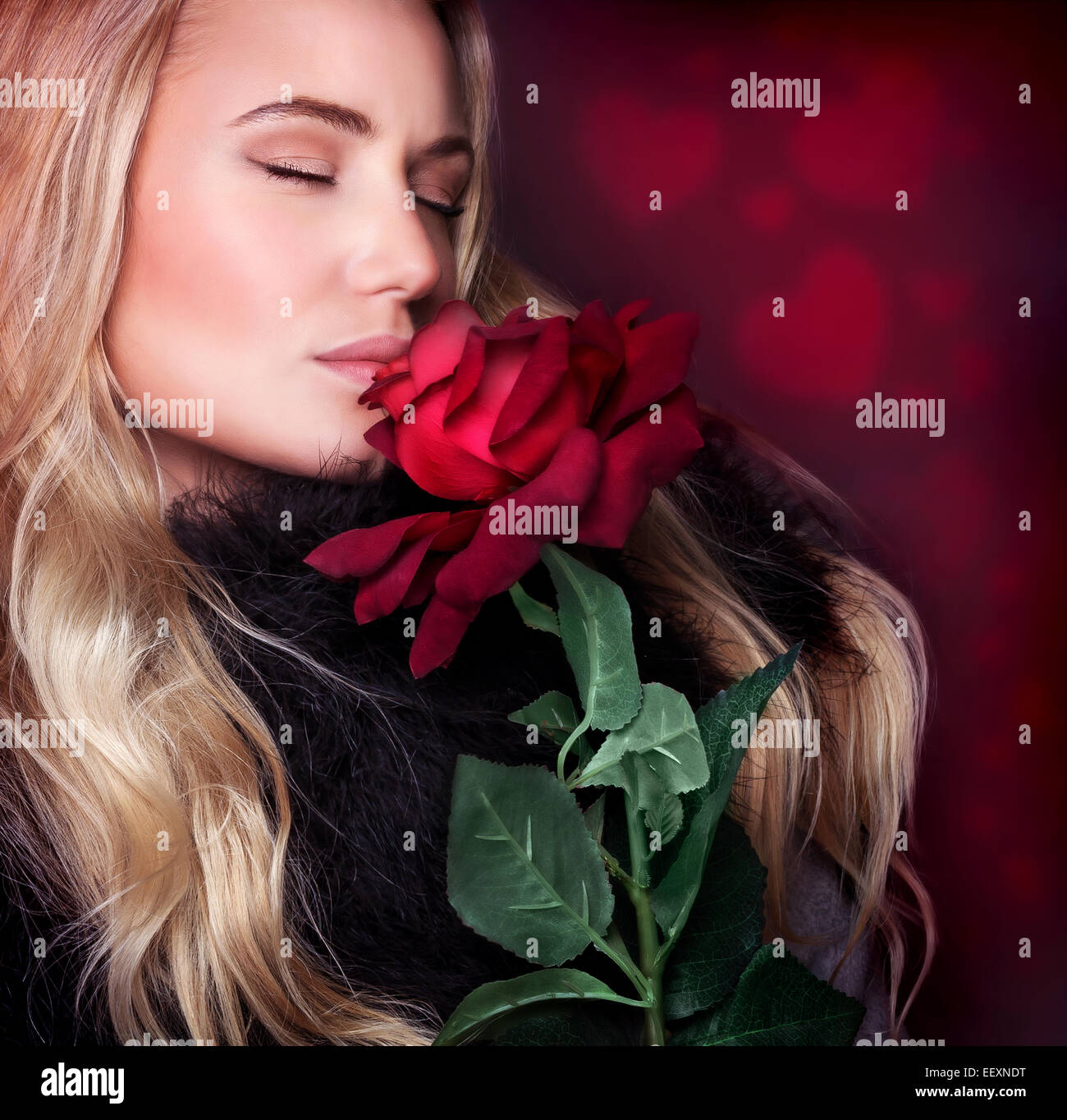 Closeup portrait of beautiful blond woman with closed eyes smelling fresh red rose on background with heart ornament Stock Photo