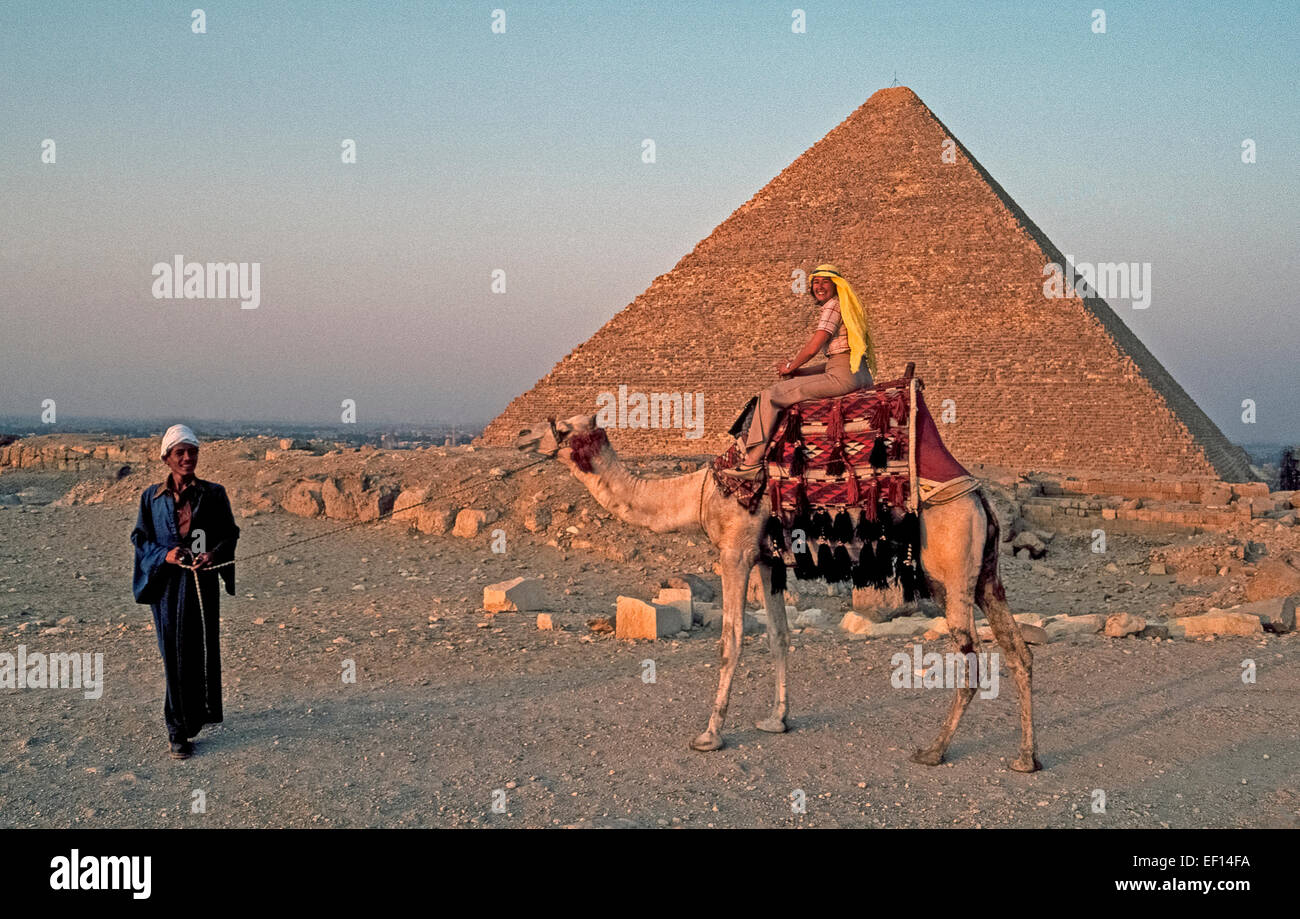A female tourist riding a camel poses for a picture in 1999 as the sun sets one of the ancient Great Pyramids at Giza on the outskirts of Cairo, Egypt. Nearly15 million tourists visited this North African nation in the year prior to the Egyptian Revolution of 2011, which has reduced that number by 45% because of subsequent turmoil in the country. Stock Photo