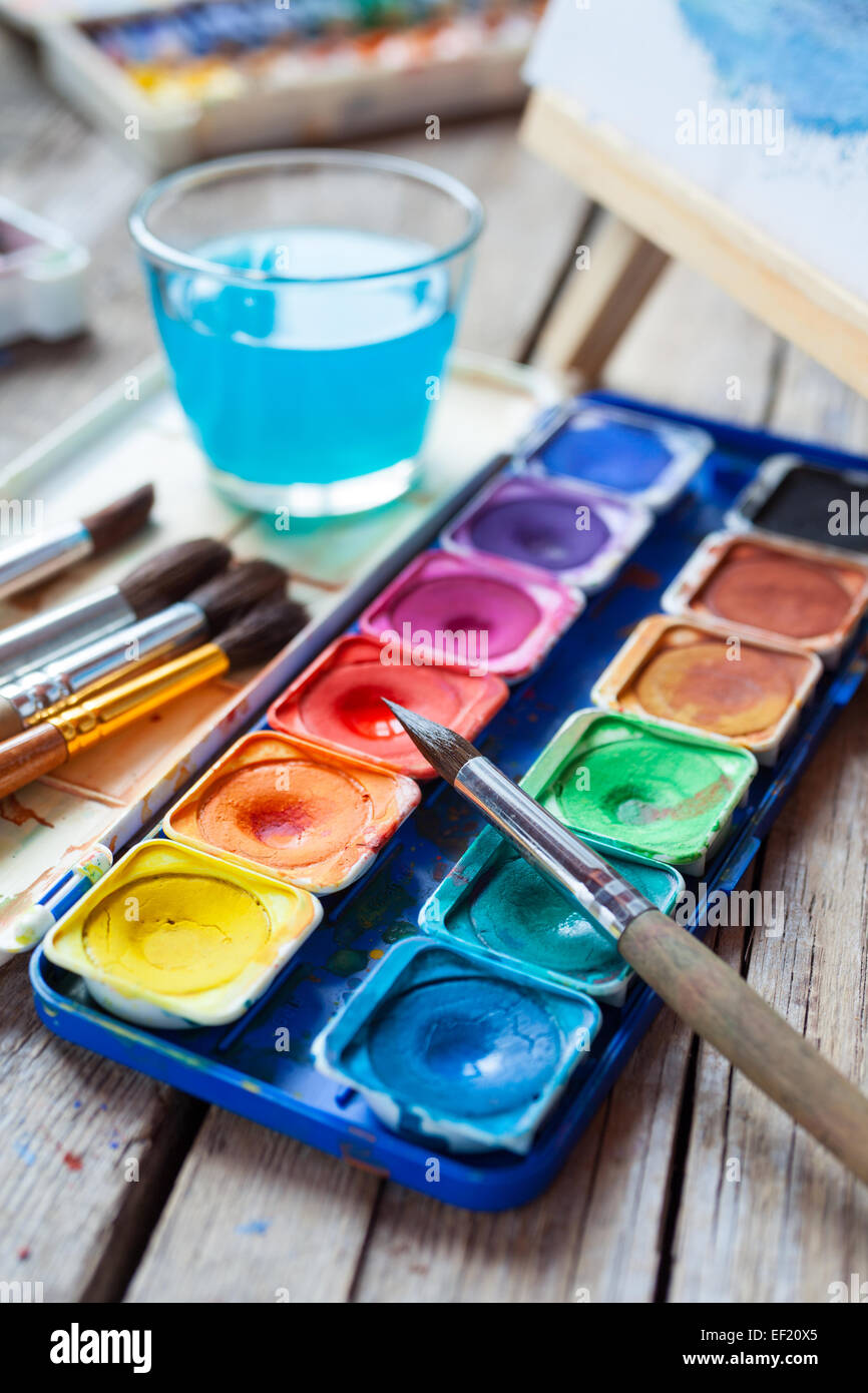 Set of watercolor paints,  art brushes,  glass of water and easel with painting on vintage wooden background. Stock Photo