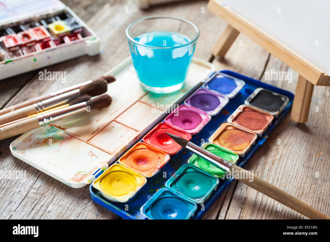 Set of watercolor paints, art brushes, glass of water and easel with painting on old wooden table. Stock Photo