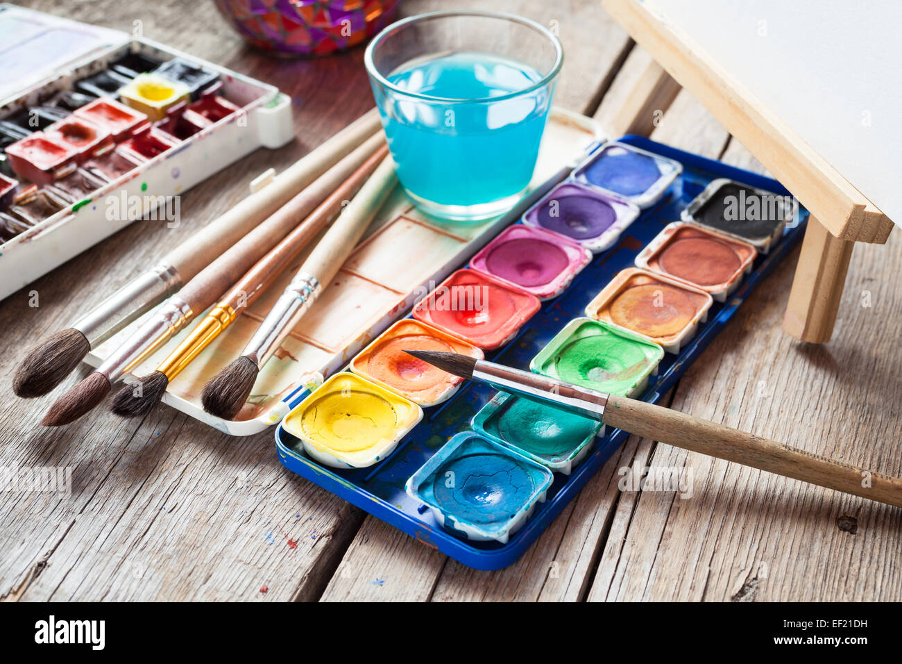 Box of watercolor paints, art brushes, glass of water and easel with canvas or paper on old wooden table. Stock Photo