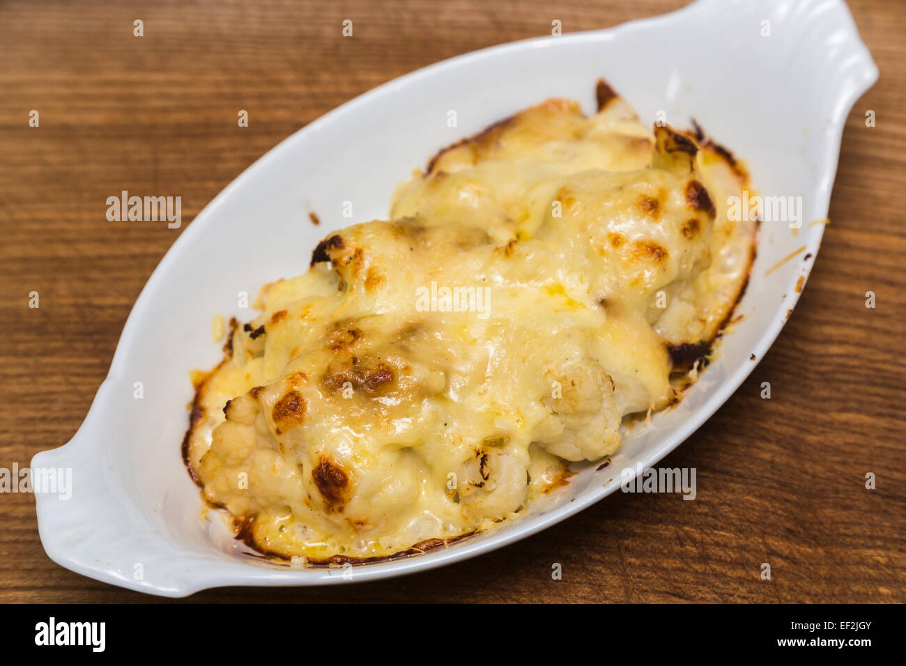 Plate of delicious home made baked cauliflower cheese served in an oval plain white china dish on a wooden board Stock Photo