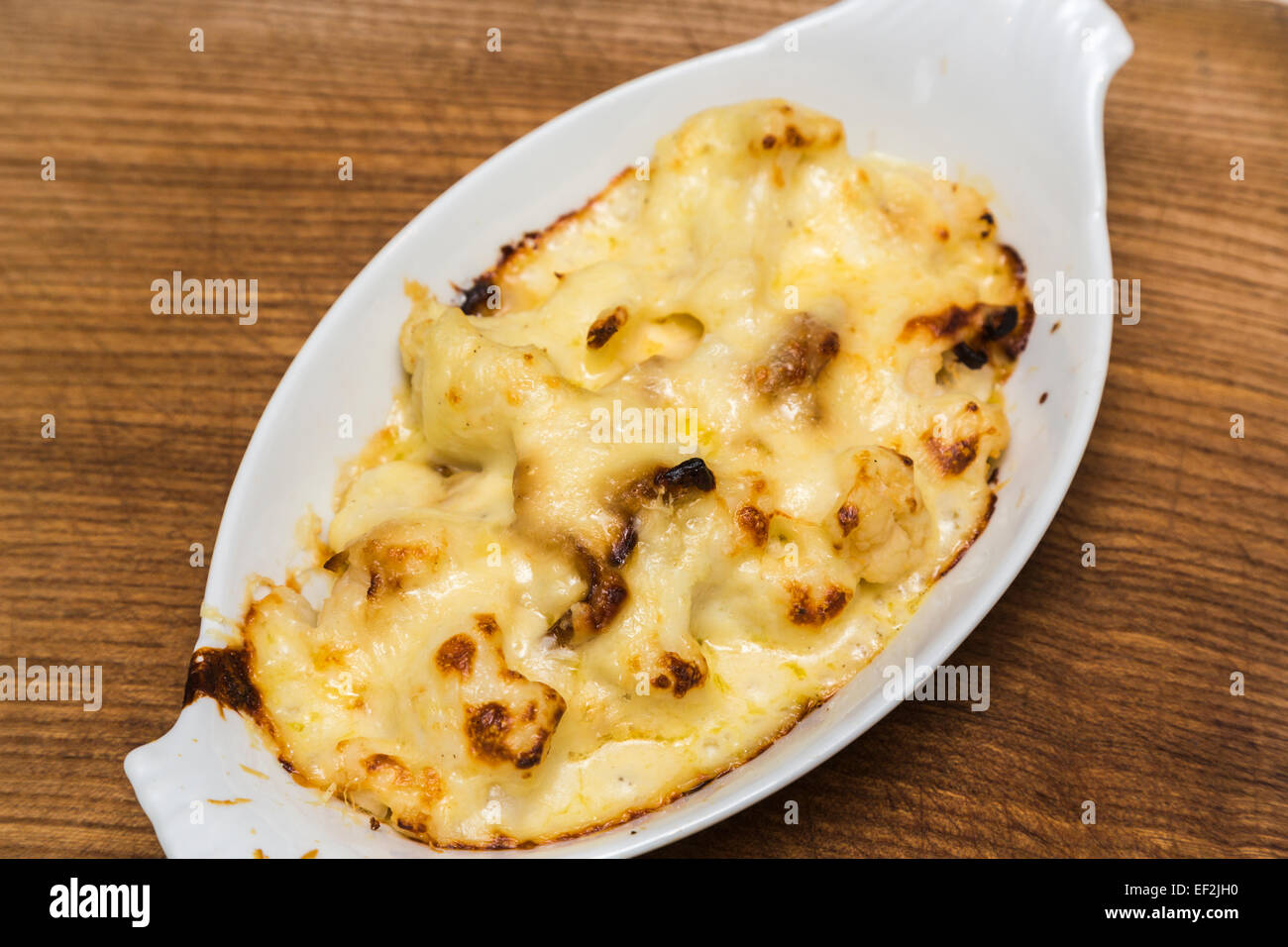 Plate of delicious home made baked cauliflower cheese served in an oval plain white china dish on a wooden board Stock Photo