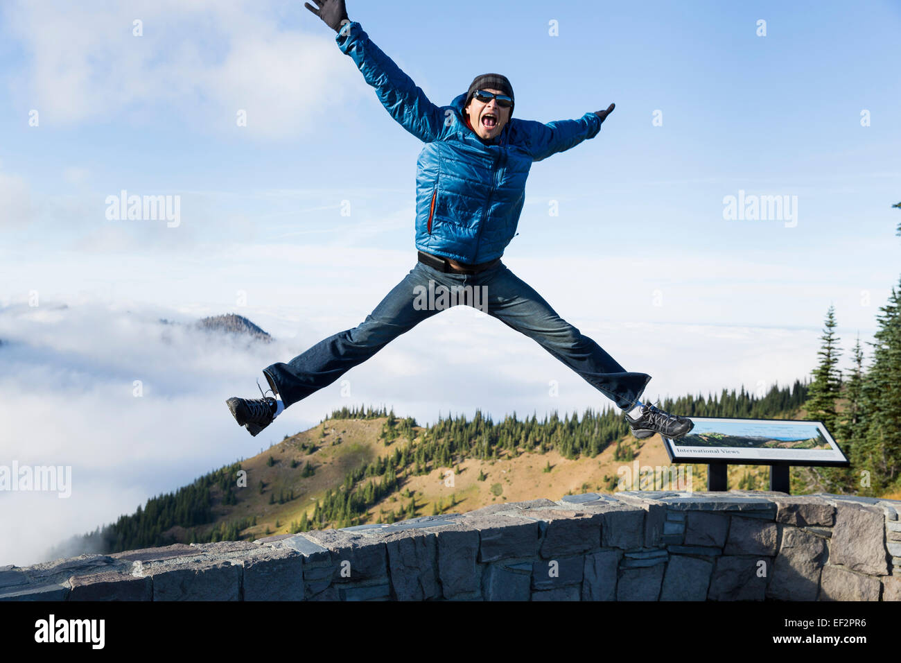 Young male jumping straight on of a wall, with his legs spread out in a funny pose, with a beautiful mountain background Stock Photo