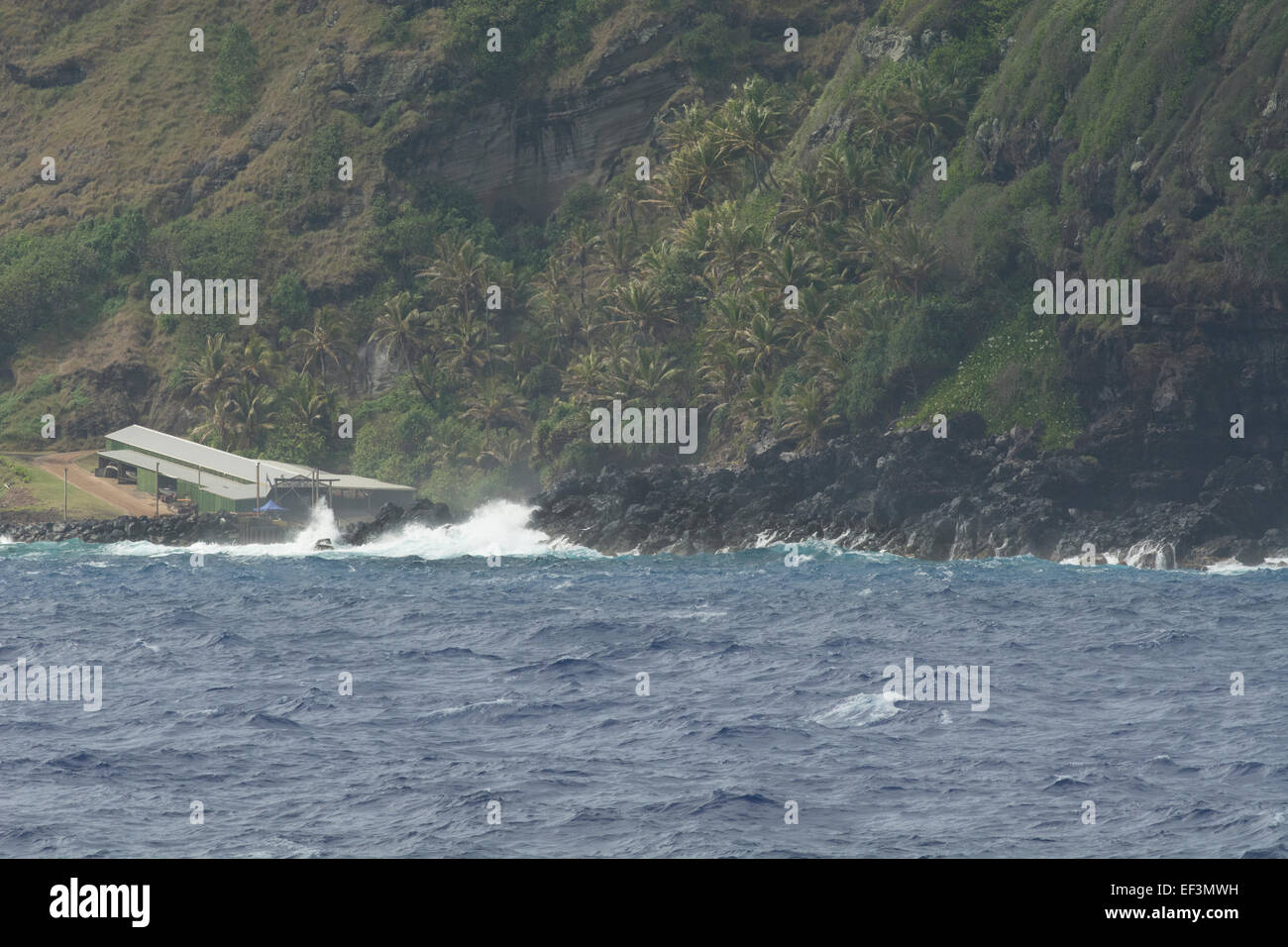 Pitcairn Islands, Pitcairn. Coastal view of rugged volcanic shore. Crashing waves around the boathouse (L) and landing site. Stock Photo
