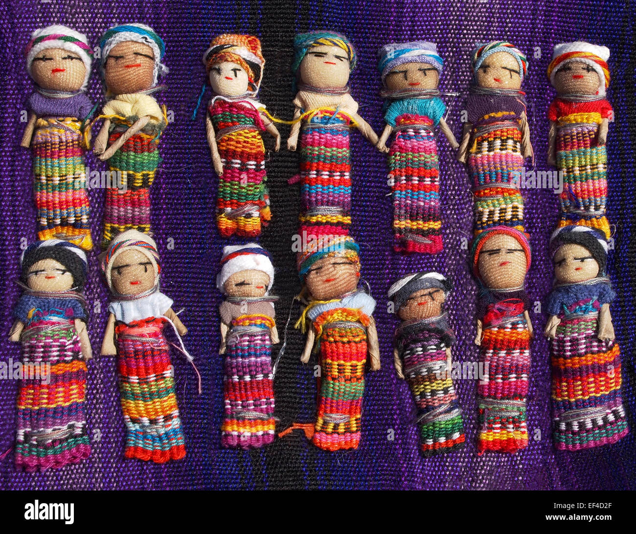 A collection of large, colorful Guatemalan Worry Dolls lined up in two rows on a purple woven blanket. Stock Photo