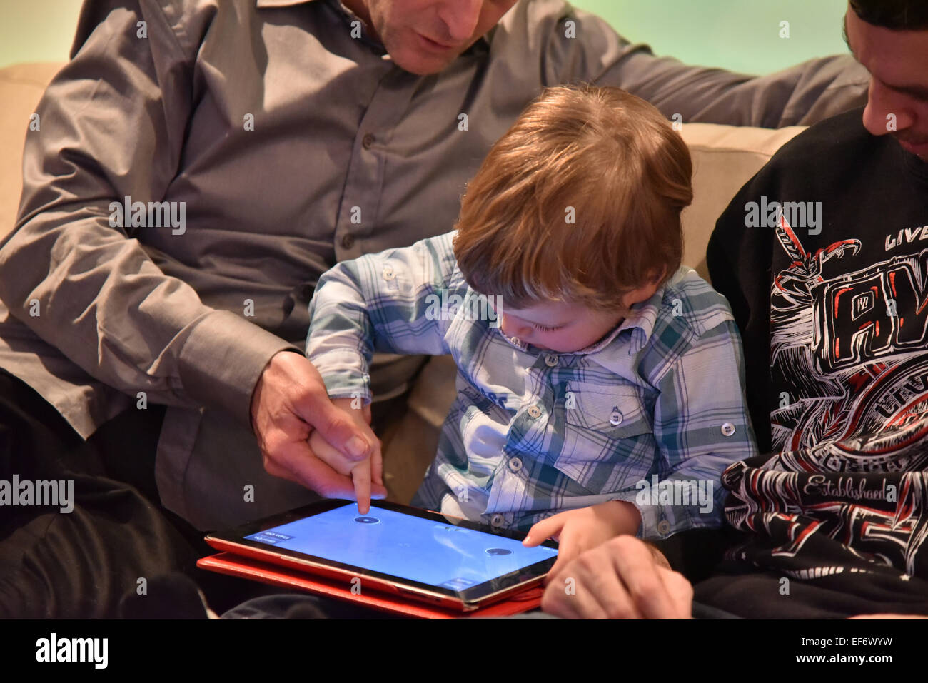 A young boy (2 1/2 yrs) playing on an iPad sat between two adults who are helping him play Stock Photo