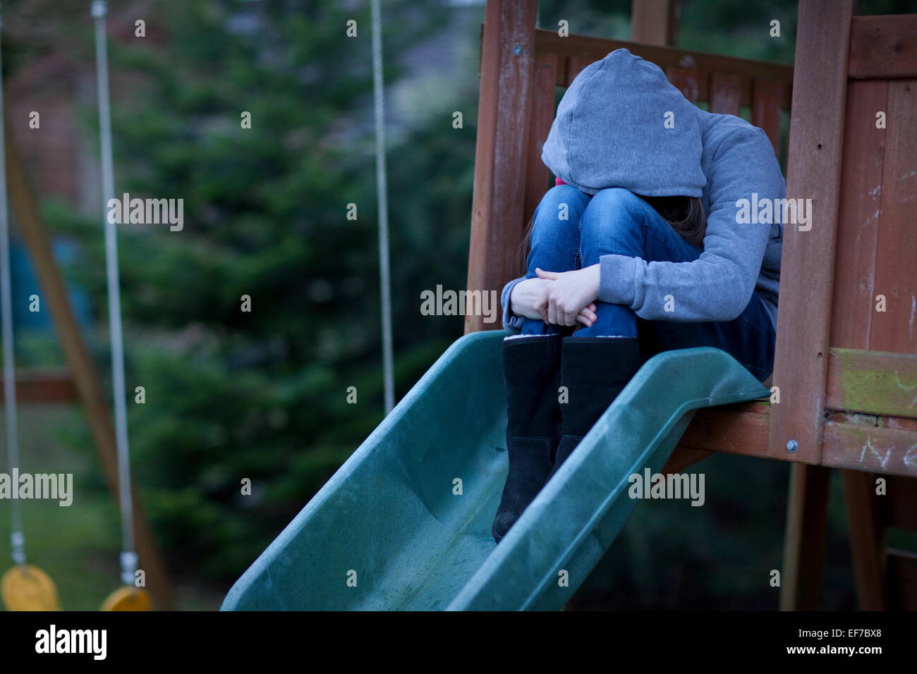 Sad child in hoodie with face hidden, sitting alone in a playground in despair. Stock Photo