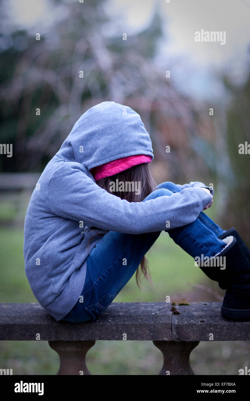Sad girl in hoodie with face hidden, sitting alone on a wall in despair. Stock Photo