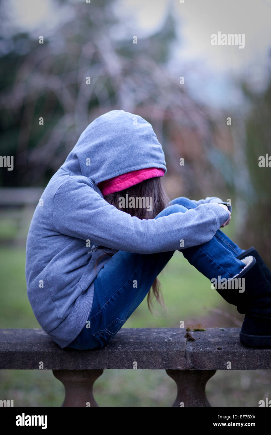 Sad girl in hoodie with face hidden, sitting alone on a wall in ...